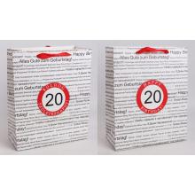 PAPIEROVÁ TAŠKA S MATNOU KLOPOU SO STUHOU, ČIERNA NA BIELOM POZADÍ, S NÁPISOM "HAPPY BIRTHDAY" V NIEKOĽKÝCH MEDZINÁRODNÝCH JAZYKOCH NA POZADÍ, ČERVENÝ KRÚŽOK S LETOPOČTOM "20"