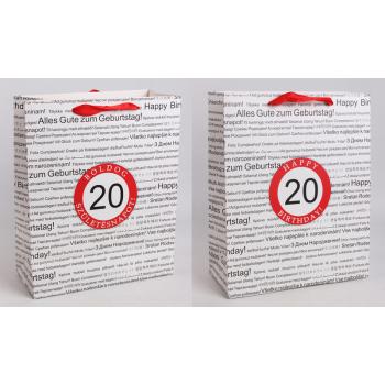 PAPIEROVÁ TAŠKA S MATNOU KLOPOU SO STUHOU, ČIERNA NA BIELOM POZADÍ, S NÁPISOM "HAPPY BIRTHDAY" V NIEKOĽKÝCH MEDZINÁRODNÝCH JAZYKOCH NA POZADÍ, ČERVENÝ KRÚŽOK S LETOPOČTOM "20"