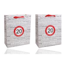 PAPIEROVÁ TAŠKA S MATNOU KLOPOU SO STUHOU, ČIERNA NA BIELOM POZADÍ, S NÁPISOM "HAPPY BIRTHDAY" V NIEKOĽKÝCH MEDZINÁRODNÝCH JAZYKOCH NA POZADÍ, ČERVENÝ KRÚŽOK S LETOPOČTOM "20"